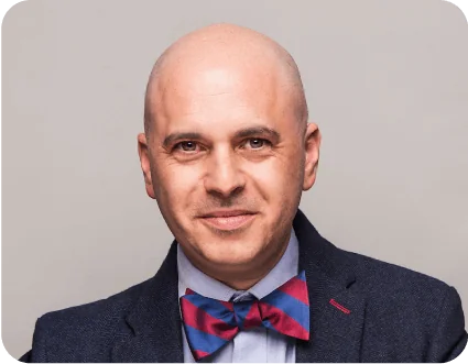 Person with shaved head smiles slightly, facing camera; wearing navy blazer, light shirt and red-and-blue striped bow tie; neutral light-gray studio...