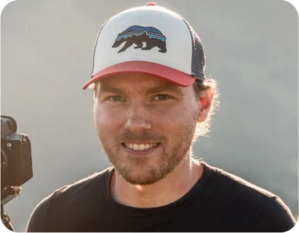 Man wearing a trucker cap with a stylized bear graphic smiles toward the camera, a mirrorless camera visible at his shoulder, warm backlit outdoor...