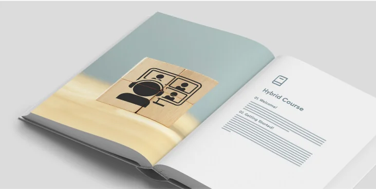 Wooden block printed with a video‑conference icon sits on an open booklet; the right page shows a "Hybrid Course" layout with section lines, all on a...