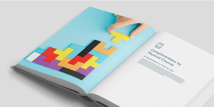 Colorful Tetris-like blocks being assembled as a hand places a yellow piece on a blue page of an open booklet on a white surface, beside a right page...