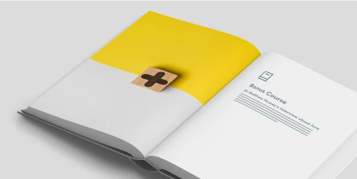 Open hardcover book lying on a light surface; left page partly yellow with a wooden block showing a black plus sign, right page displaying a minimal...