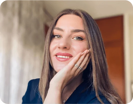 Young woman smiling, resting her chin on her hand, showing rings; long straight hair and makeup; seated indoors with soft light, blurred curtains and...