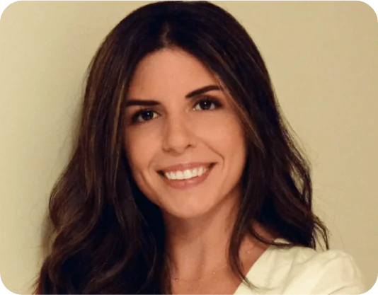Woman with long dark hair smiles directly at the camera, wearing a white blouse against a plain beige wall under soft, warm indoor lighting.