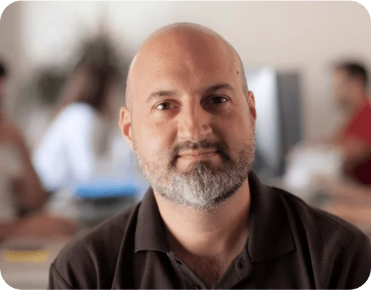 Bald man with short gray beard gazes directly at camera, seated and slightly smiling, wearing a dark polo; blurred coworkers and computer monitors...