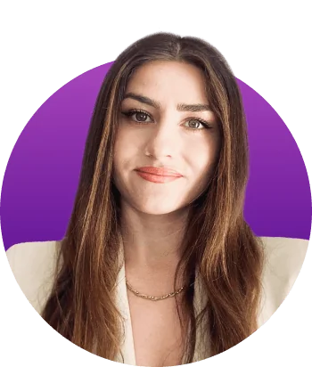Woman smiles slightly, facing the camera; wearing a cream blazer and gold chain, long brown hair framing her face, arched eyebrows and subtle makeup...