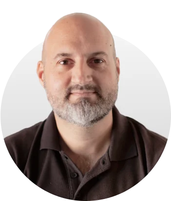 Adult with shaved head and short gray beard faces camera, wearing a dark polo shirt; portrait centered in a soft, light-gray circular frame against a...