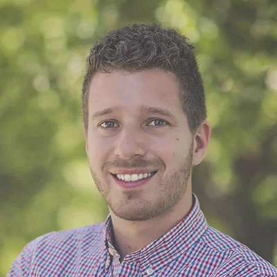 Young man smiles directly at the camera, head-and-shoulders pose; short curly hair, light stubble, wearing a red-and-blue checked shirt; blurred...