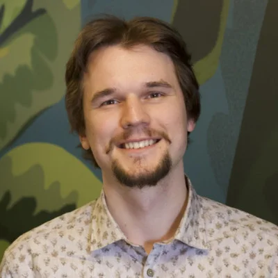Young man in his late 20s smiling directly at the camera; wearing a patterned button-up shirt and standing in front of a painted green, leafy mural...