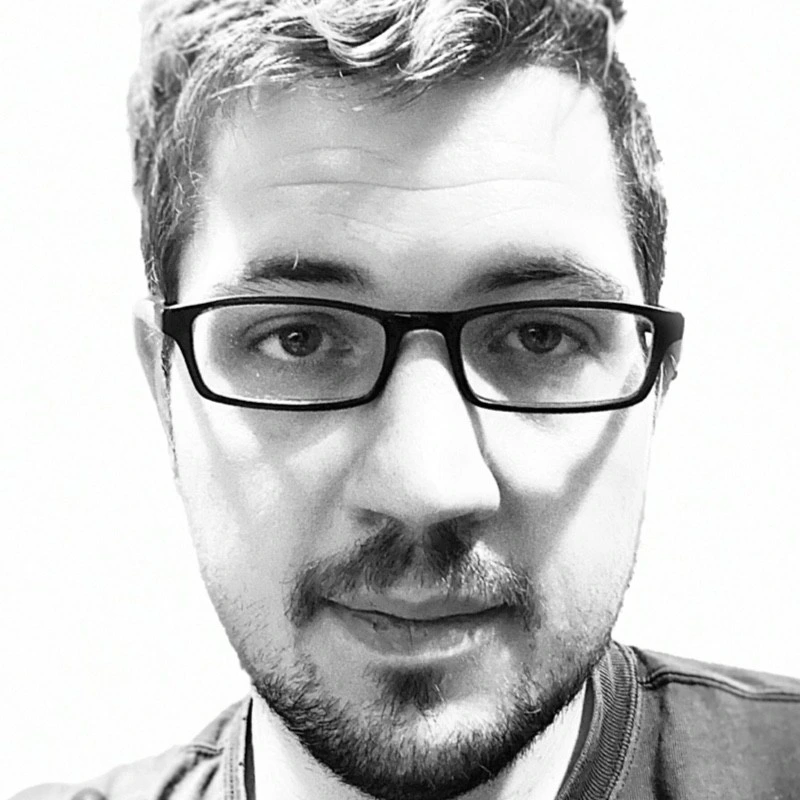Young bearded man wearing rectangular glasses looking directly at camera in a high-contrast black-and-white close-up portrait against a plain light...