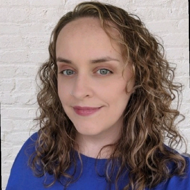 Person with shoulder-length curly light-brown hair, wearing a blue top, smiling slightly and facing the camera; white painted brick wall behind them.