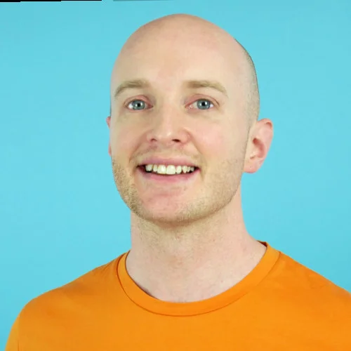 Bald man with light stubble smiles at the camera, wearing an orange T-shirt, posed against a solid light-blue studio background.