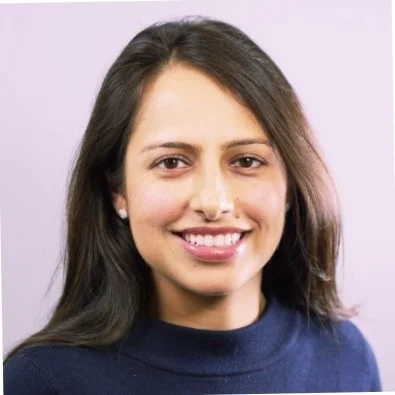 A young woman with long dark hair smiles directly at the camera, wearing a navy sweater and small pearl earrings, against a soft light-purple...