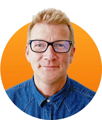A middle-aged man wearing black-framed glasses and a buttoned denim shirt looks directly at the camera with a slight smile, set against a bright...