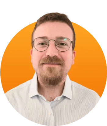 Head-and-shoulders portrait of a bearded man wearing round glasses and a light-striped shirt, looking at the camera with a slight smile against an...