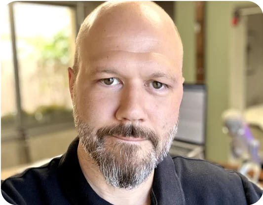 Adult with bald head and trimmed beard looks directly at camera, seated in a home office with blurred window, laptop on desk and microphone visible...