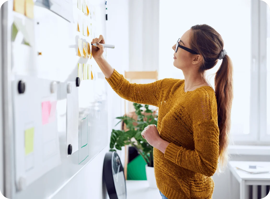 Woman in a mustard sweater writes on sticky notes attached to a whiteboard, standing in a bright office near a window, desk, potted plant and wall...