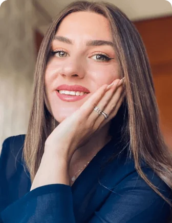 Young woman smiles, resting her chin on one hand showing two rings; long straight hair and light makeup; wearing a dark blazer in a softly lit indoor...