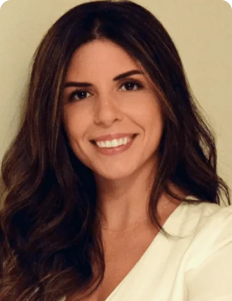 A woman with long wavy brown hair smiles at the camera, wearing a white blouse against a softly lit neutral beige background, presenting a relaxed,...