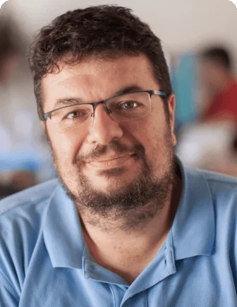 Bearded man in rectangular glasses smiles gently at the camera, seated indoors with soft, blurred background figures; wearing a light blue polo shirt...