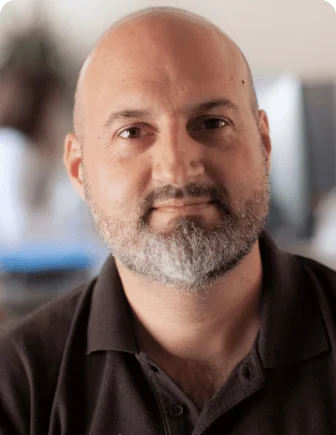 A bald man with a gray beard gazes directly at the camera with a calm expression while seated in a softly lit office with blurred monitors and plant...