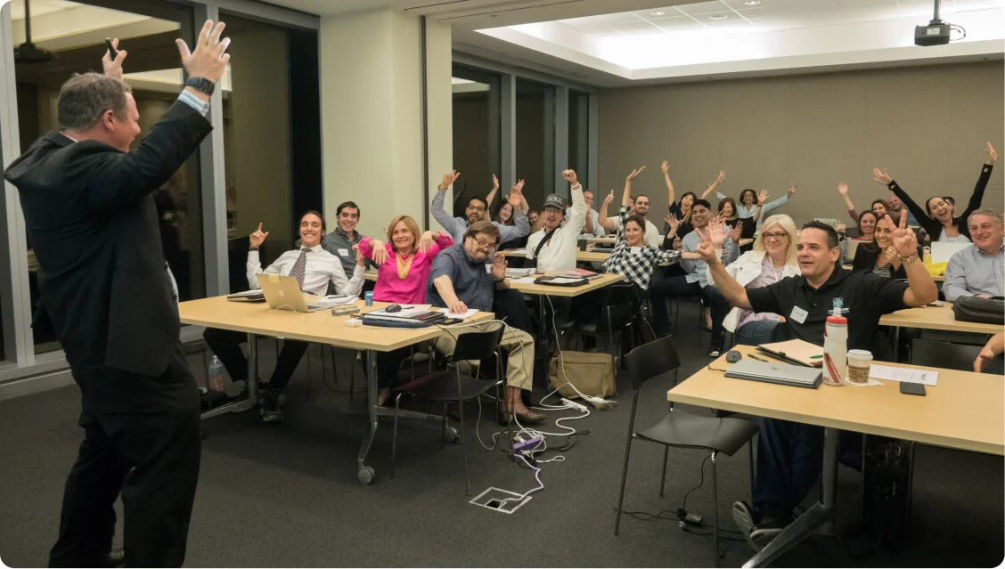 Audience raising hands toward a standing presenter in a conference-style classroom, smiling and cheering while seated at tables with laptops,...