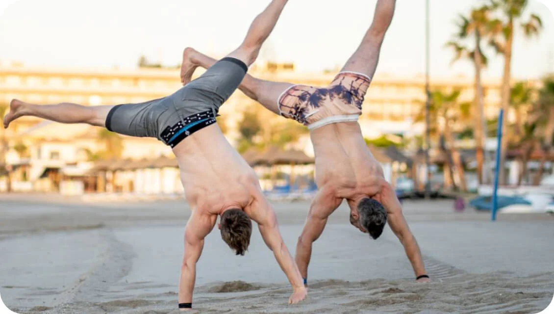 Two shirtless men do simultaneous cartwheels on a sandy beach, legs extended mid-air; resort buildings, palm trees, and beach gear blur in warm...