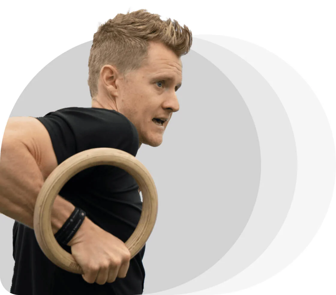 Male athlete gripping a wooden gymnastic ring, pulling himself upward with tense arm muscles; focused expression, black shirt against a minimalist...