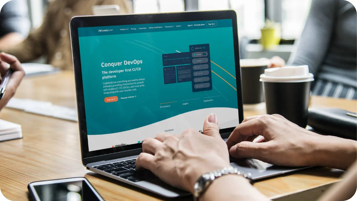 Laptop screen shows a Codefresh webpage promoting DevOps while hands type on the keyboard at a wooden meeting table with coffee cups and a phone...