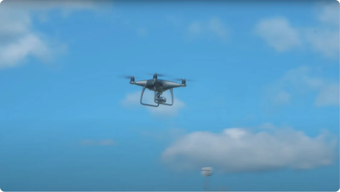 A quadcopter drone hovers midair with its camera gimbal visible, framed against a bright blue sky with scattered white clouds; a blurred light pole...