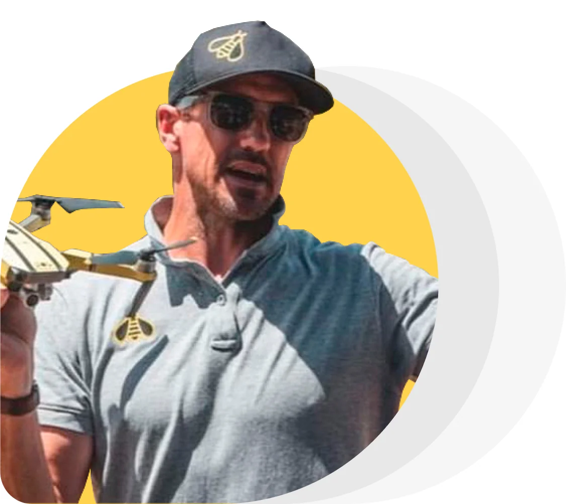 Man wearing sunglasses and a baseball cap with a bee emblem holds a small quadcopter drone at chest level, speaking, against a yellow circular...