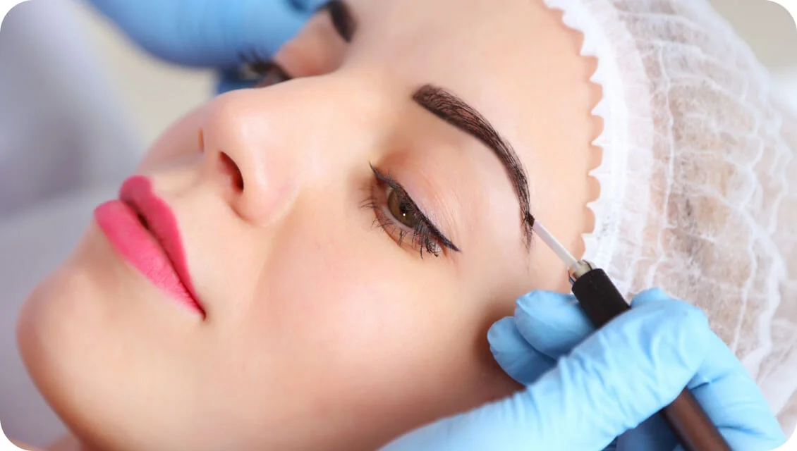 Eyebrow receiving microblading: a gloved technician uses a handheld tool to apply pigment while the client reclines in a clinical setting wearing a...