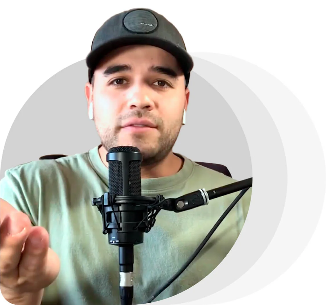 Person wearing a dark baseball cap and green shirt gestures while speaking into a studio microphone; white wireless earbuds visible against a neutral...