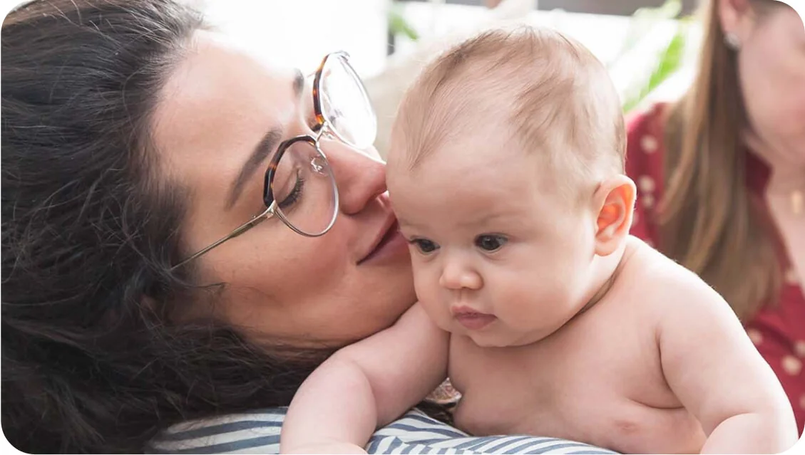 Woman cradles and nuzzles a shirtless infant who gazes downward, both on a striped cushion in a softly lit indoor room with a blurred adult seated in...