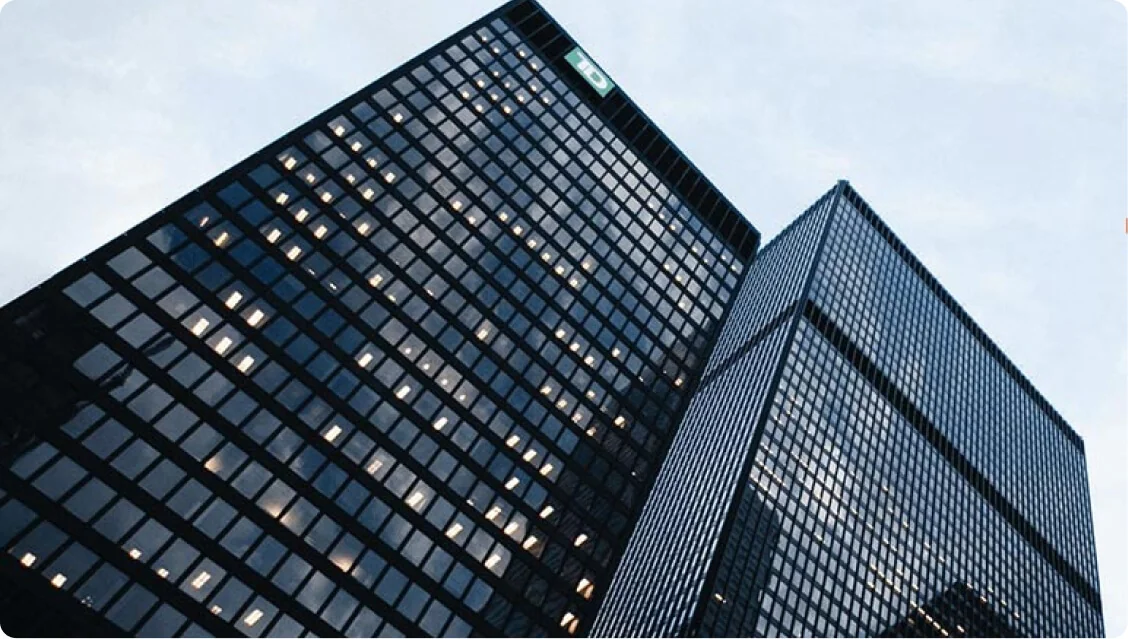 Two glass-clad skyscrapers tilt upward, their gridlike windows reflecting a pale sky while many interior lights glow; viewed from street level...