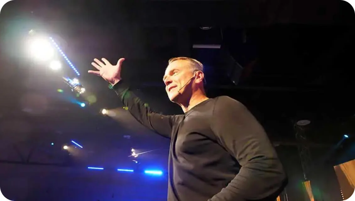 A man wearing a headset microphone raises his arm and gestures toward the audience while standing on a dim stage illuminated by bright spotlights and...