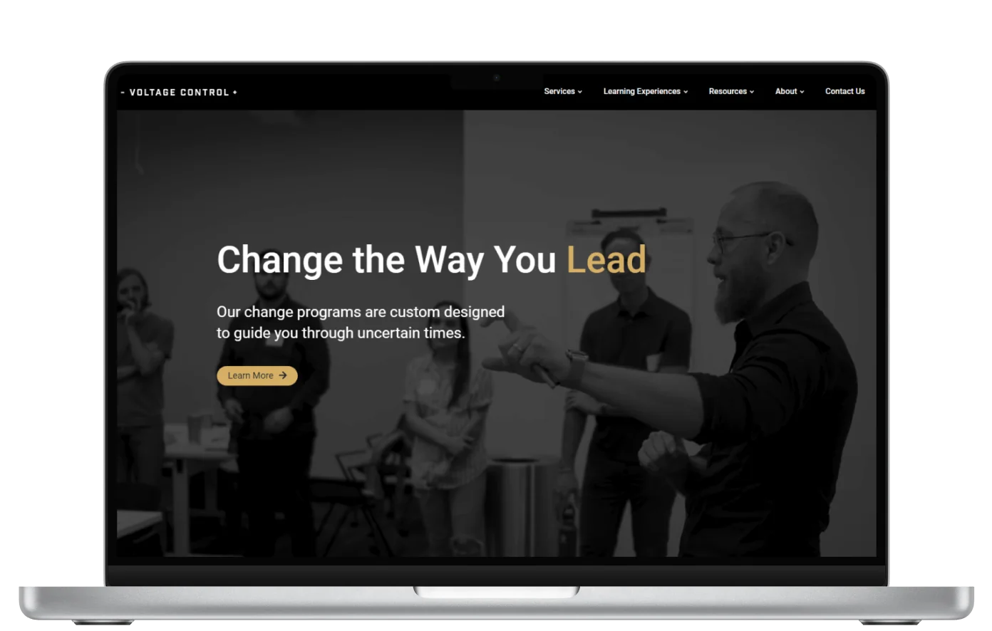 Website headline overlays a grayscale photo of a bearded man gesturing to a small group in an office, shown on a laptop screen.