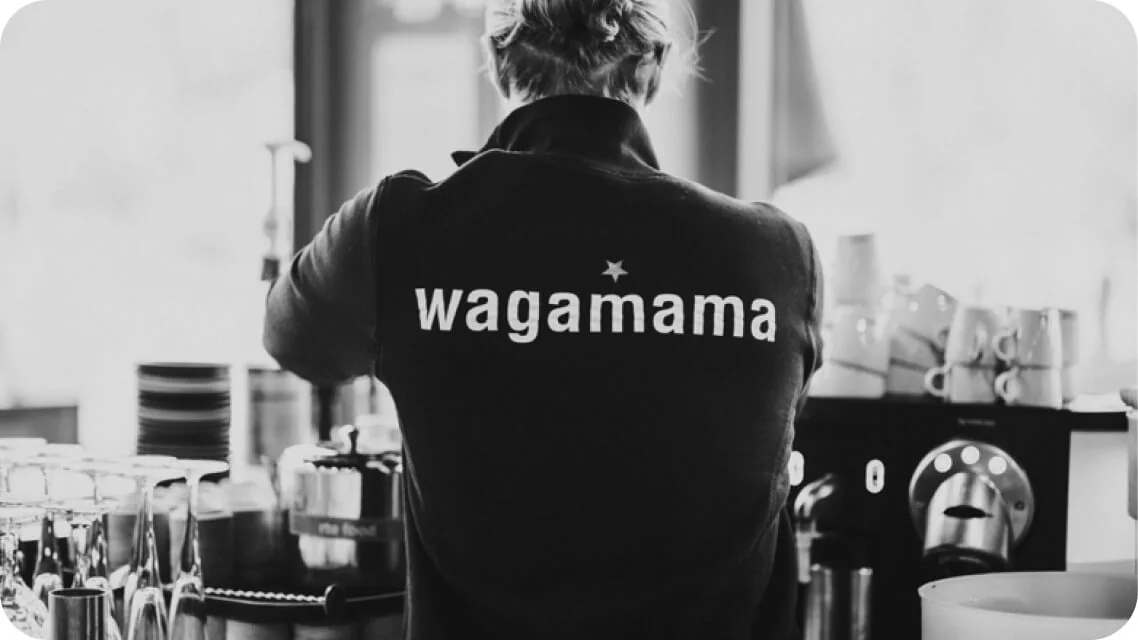 Staff member (rear view) wearing a "wagamama" jacket, operating an espresso machine and arranging cups amid glassware and stacked dishes at a bright...