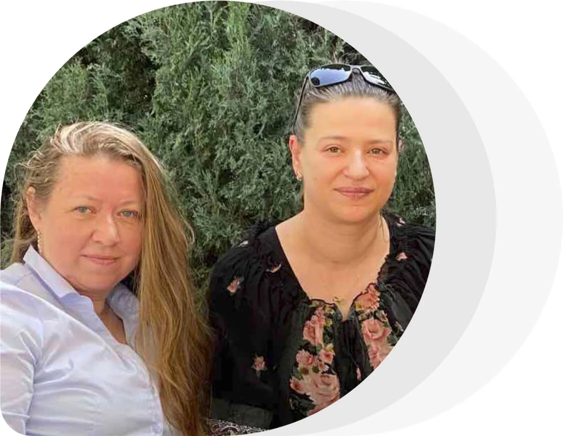 Two women sit close, facing the camera with smiles; one wears a white shirt and long hair, the other a floral blouse with sunglasses atop her head,...