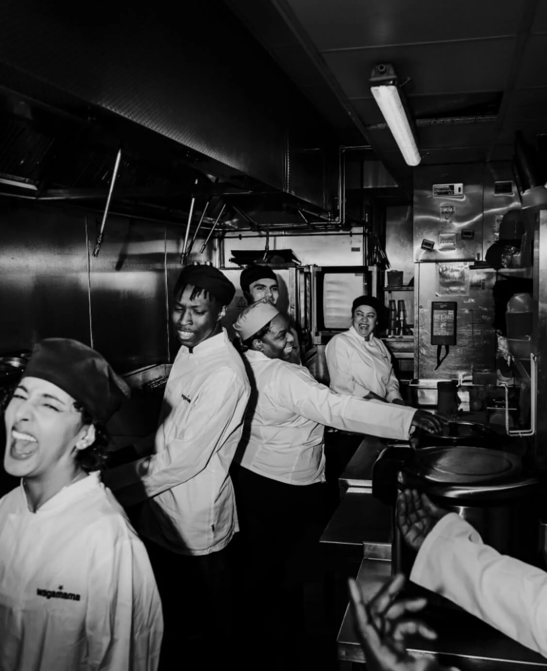 Group of five chefs laugh and reach for pots while cooking in a narrow stainless-steel commercial kitchen, working shoulder-to-shoulder beneath a...