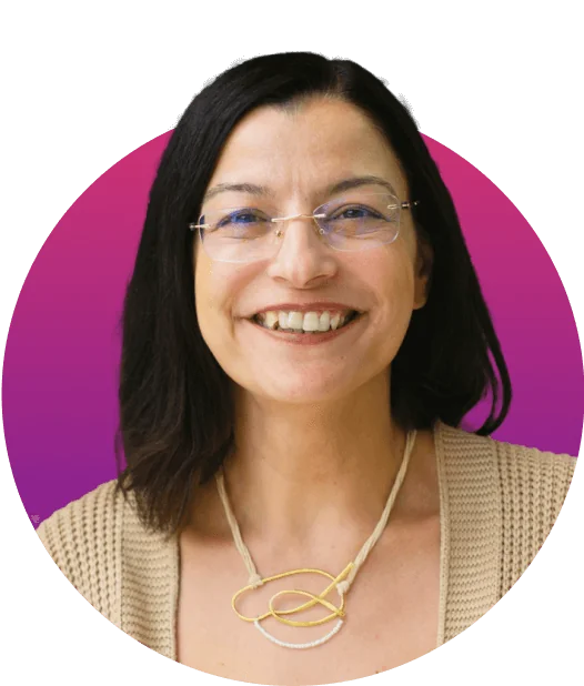 Smiling woman wearing glasses and a beige cardigan, displaying a gold abstract necklace, facing the camera; short dark hair against a circular...