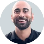 Bald man with trimmed beard smiling directly at the camera, wearing a dark polo shirt; centered against a pale gray circular background.