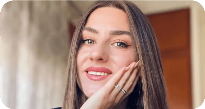 A smiling woman rests her chin on her hand, showing rings; she faces the camera in a close-up, softly lit indoor setting with curtains and a wooden...