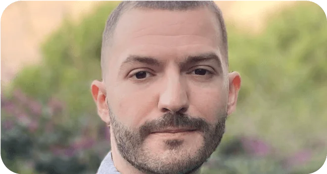 A bearded man looks calmly at the camera; soft-focus garden greenery and purple flowers form the blurred background behind him.