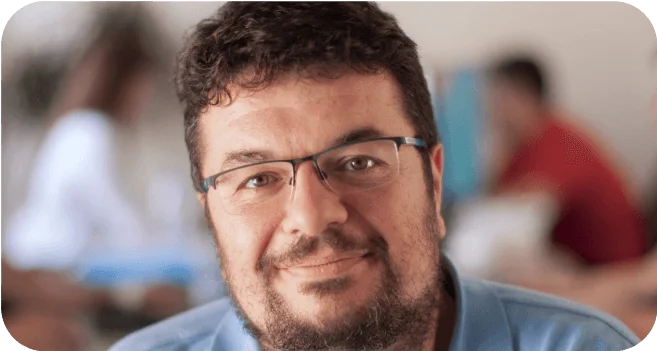 Bearded man wearing glasses and a light blue shirt smiles gently at the camera, seated in a softly lit indoor space with blurred figures and colorful...
