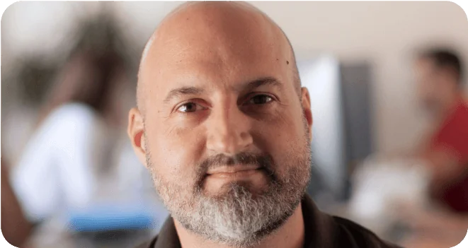 Bald bearded man looking directly at the camera, softly smiling; seated in a blurred office environment with coworkers and desks visible in the...