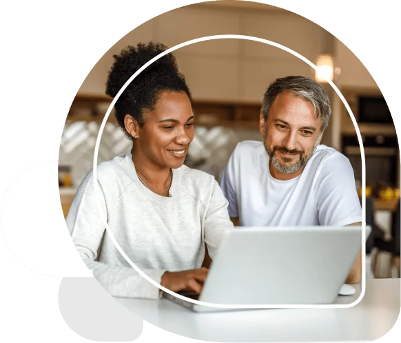 A woman and a man look at a laptop together, smiling, while seated at a kitchen counter with warm lighting, blurred cabinets and a pendant light...