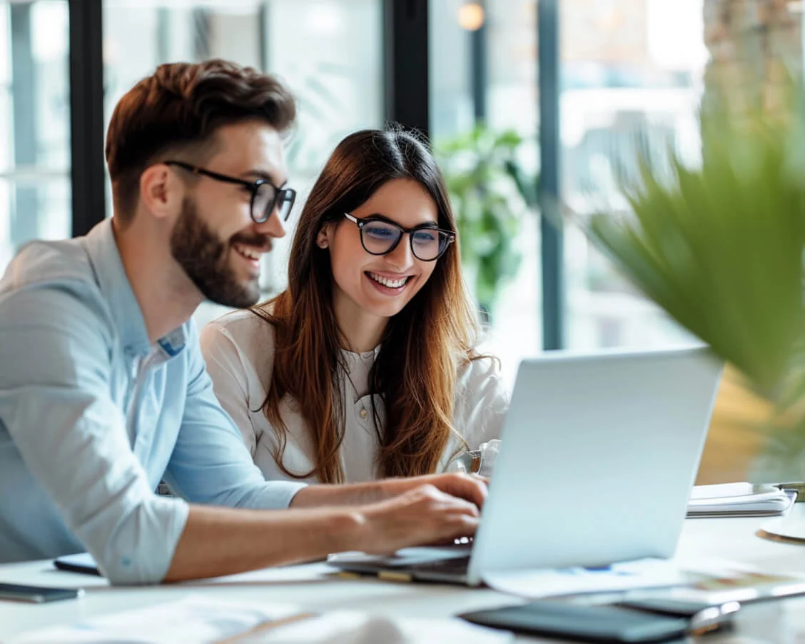 Two colleagues, a man and a woman, smile and type on a laptop while sitting at a sunlit office desk with papers, a smartphone, and potted plants near...