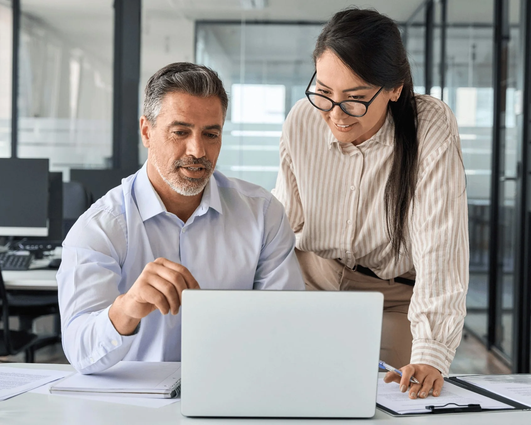 A man and woman review a laptop screen, the man pointing while the woman leans over; office with glass partitions, desks, papers, notebooks and a...