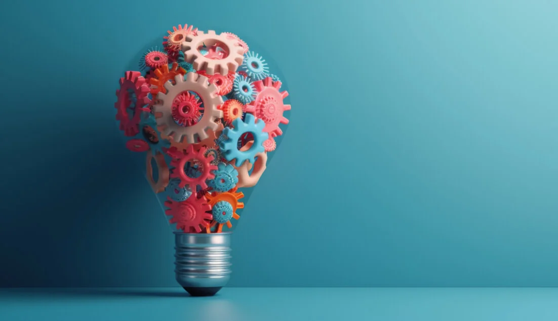 Lightbulb-shaped cluster of interlocking pink, red, orange and blue gears forming a bulb, standing upright on a flat surface with soft teal...