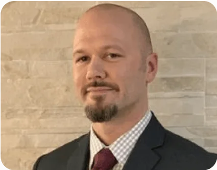 Bald man with short goatee wearing a dark suit, checked shirt, and maroon tie, posing with a slight smile while facing the camera against a light...